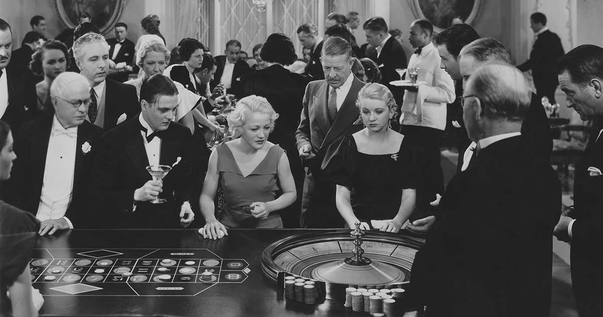 Historische Aufnahme einer dicht gedrängten Rouletteszene im Casino, elegant gekleidete Menschen beobachten das Spiel am Roulettetisch.