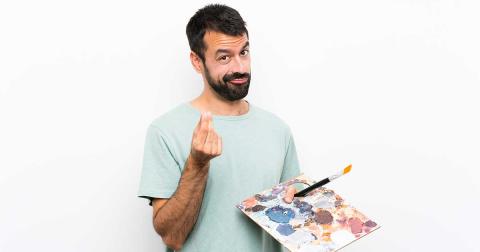 Junger Künstler hält Palette und zeigt Geld-Geste – Symbol für Finanzen in der Kulturbranche.