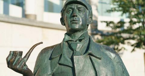 Statue einer literarischen Detektivfigur mit Pfeife in der Hand, Nahaufnahme vor unscharfem Hintergrund.